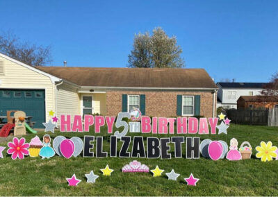 YL Hampton sign on front lawn in front of a house during the day - "Happy 5th Birthday Elizabeth". Pink, silver and black letters surrounded by similarly colored elements - balloons, princesses, flowers, stars and cupcakes.