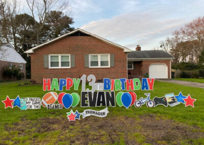 YL Hampton sign on front lawn in front of small house - "Happy 13th Birthday Evan". Multicolored red, green and blue letters ("13th" is silver, "Evan" is black), balloons, football, bike and video game elements. "I paused my game for this". "#TEENAGER".