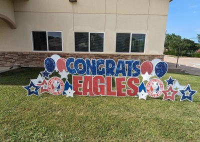 This image features a celebratory yard sign that reads "CONGRATS EAGLES" in bold, glittery blue and red letters. The sign is decorated with patriotic-themed stars, balloons, and two circular emblems displaying the "Eagle Scout" logo of the Boy Scouts of America. The setup is placed on a well-maintained grassy area in front of a beige building with stone accents and several windows. The background includes a parking lot, trees, and a clear blue sky, suggesting an outdoor daytime setting.