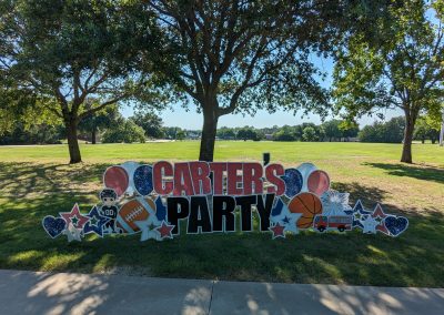A large, colorful yard sign displayed in a park-like setting with green grass, trees, and a clear blue sky. The sign reads 'Carter's Party' in bold red and black letters. Surrounding the text are various themed decorations, including balloons, stars, a hockey player, a football, a basketball, a fire truck, and fireworks. The festive sign is set against an open field with trees providing shade, suggesting an outdoor celebration.