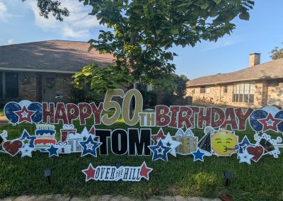 A large, colorful yard sign displayed on a well-maintained lawn in front of a brick house with a tree and bushes. The sign reads 'Happy 50th Birthday Tom' in bold, decorative letters, with '50' emphasized in gold. The design includes various celebratory elements such as stars, balloons, a birthday cake, presents, and a smiling emoji blowing a party horn. At the bottom, a smaller sign reads 'Over the Hill,' indicating a humorous take on the milestone birthday.