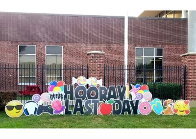 Lawn Sign in front of School: Hooray Last Day (Black); Lawn Graphics: Happy face with shades, head phones, game controller, lollipops, Ring Pop, disco dancer shadows, balloons, wrapped multi-colored round candies, Donuts, flip-flops, puppy, happy face with haearts, pencil, and wind spinner - pinks, blue, red, yellow and teal - also an apple front and center.
