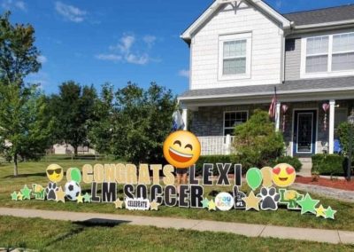 YL Little Miami sign - "Congrats Lexi LM Soccer". Gold and black letters surrounded by green balloons and stars, LM school letters, soccer ball and pawprints.