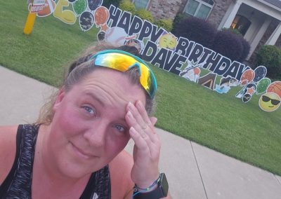 YL Emerald Coast sign - "Happy Birthday Dave". Bold black text surrounded by black, orange and green glittery balloons, emojis, sports, hunting and fishing symbols. A woman wearing sunglasses poses in front of the sign with a hand on her forehead.