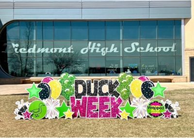 Piedmont High School in the background with Piedmont High School on a wall of windows. Lawn Sign Letters: Duck (black), Week (Bright Pink); Lawn Sign Graphics: Green duck in circle, Stars, Balloons and fireworks (green, black yellow pinkm), Wrapped 20 years in a cirlcle and celebration megaphones with streamers.