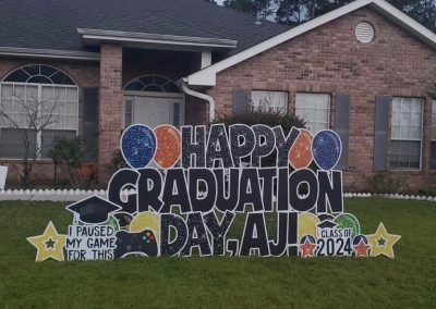 YL Pensacola sign - "Happy Graduation Day, AJ!". Glittery black letters, orang and blue balloons and yellow stars. "I paused my game to be here". "Class of 2024".