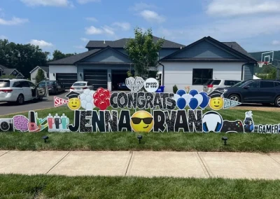 YL CT sign - "Congrats, Jenna and Ryan". Glittery black letters surrounded by red, white and blue balloon accents and emojis with graduation caps. Photography and Stanley cups for Jenna, video games and golf for Ryan